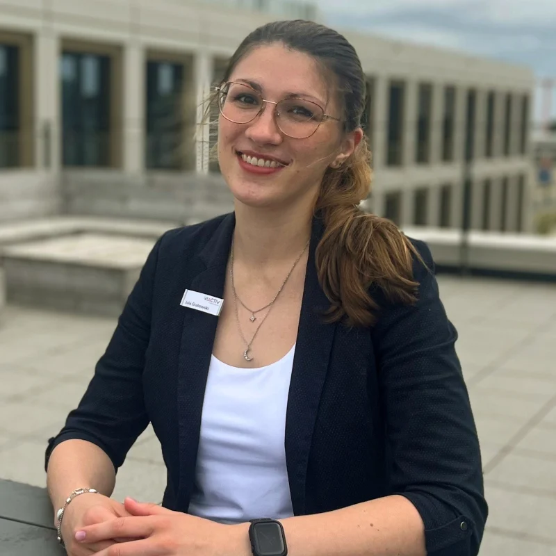 Eine Mitarbeitende der VIACTIV Krankenkasse sitzt auf einer Terrasse und lächelt in die Kamera. | VIACTIV Krankenkasse