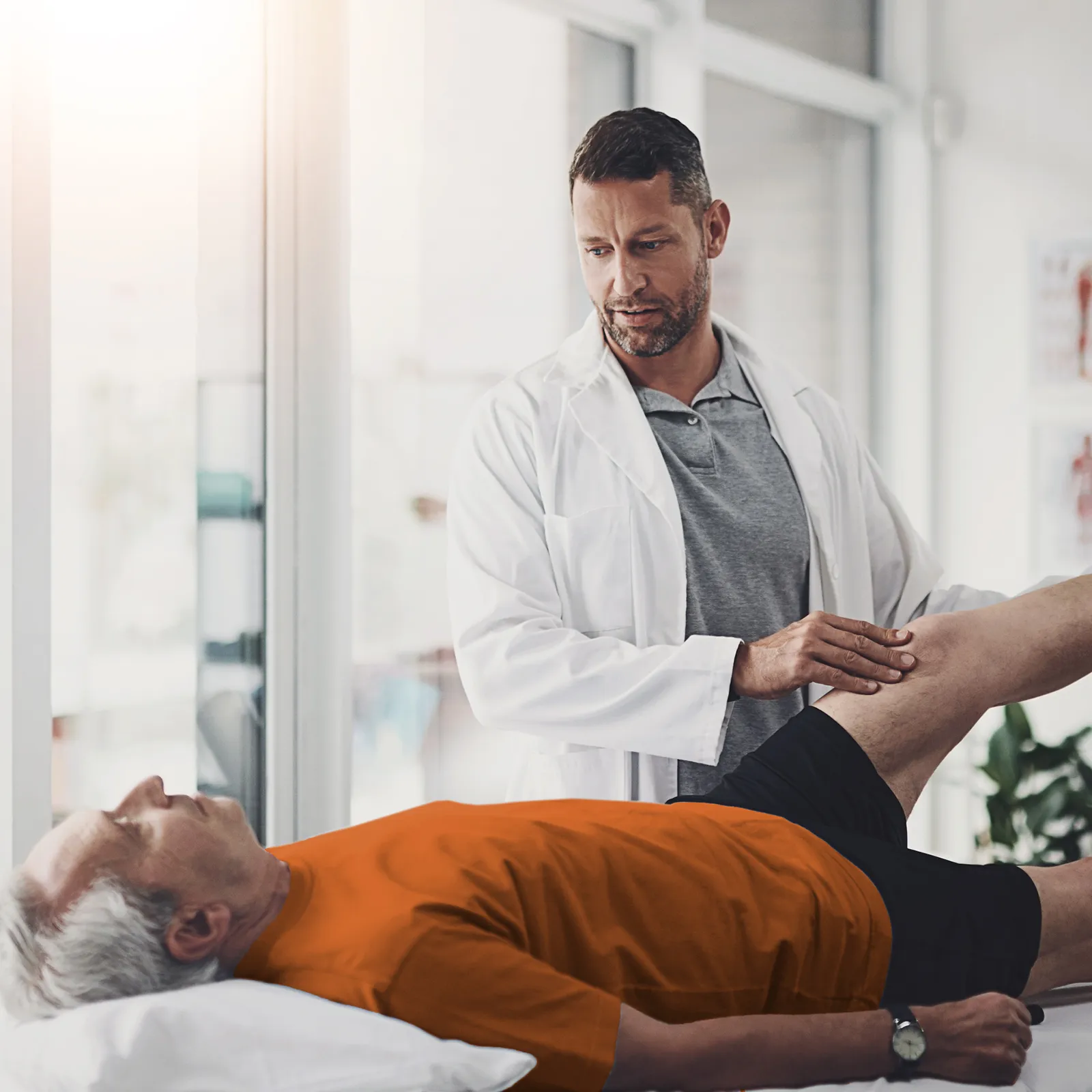 Ein Patient liegt auf einer Liege. Vor ihm steht ein Arzt, der sein Knie abtastet. | VIACTIV Krankenkasse