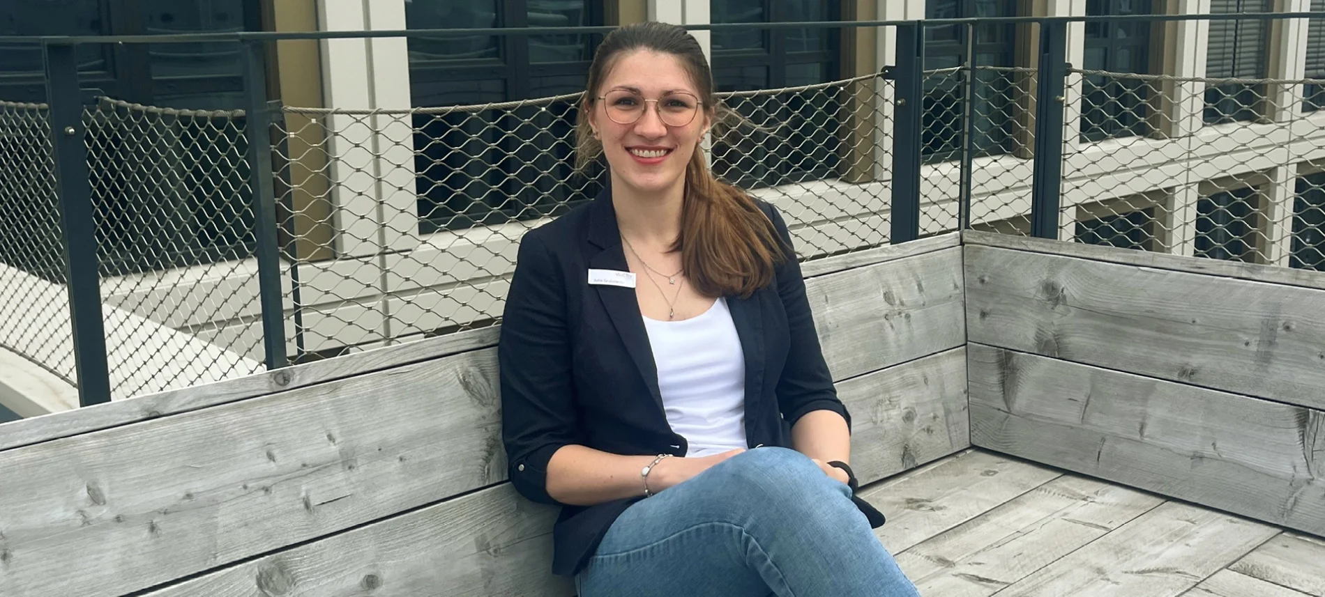 Eine Mitarbeitende der VIACTIV Krankenkasse sitzt auf einer Terrasse und lächelt in die Kamera. | VIACTIV Krankenkasse