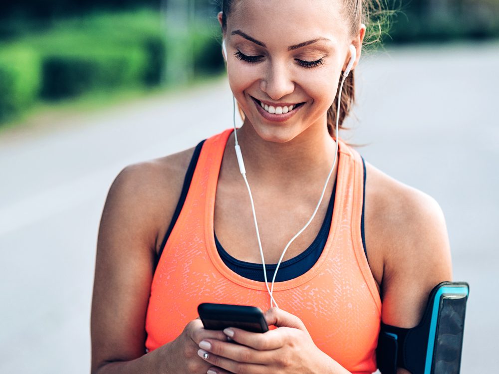 Eine Frau hat Kopfhörer in den Ohren und ist bereit joggen zu gehen. Sie startet die Musik an dem Telefon. | VIACTIV Krankenkasse
