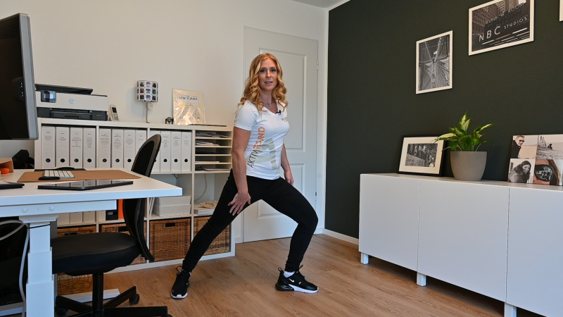 Frau im Arbeitszimmer zeigt Bewegungen zur Stärkung der Lendenwirbelsäule. | VIACTIV Krankenkasse