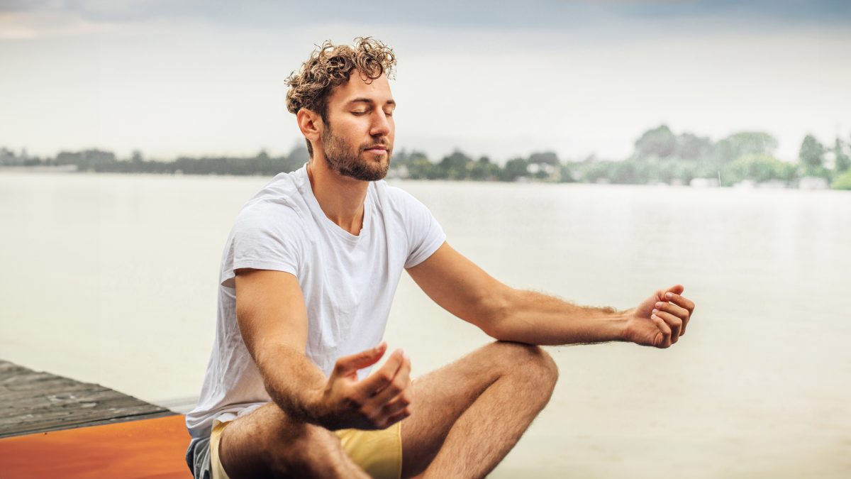 Ein junger Mann sitzt an einem See und macht Yoga. | VIACTIV Krankenkasse
