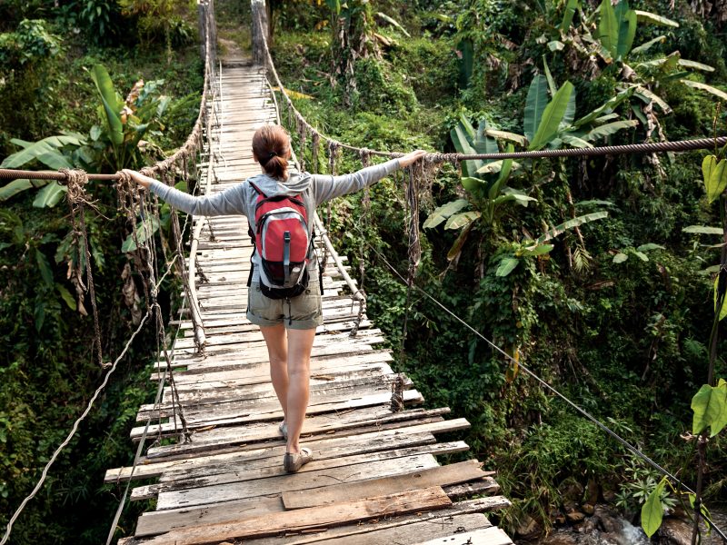 Eine junge Frau balanciert auf einer Auslandsreise vorsichtig über eine hölzerne Hängebrücke. | VIACTIV Krankenkasse