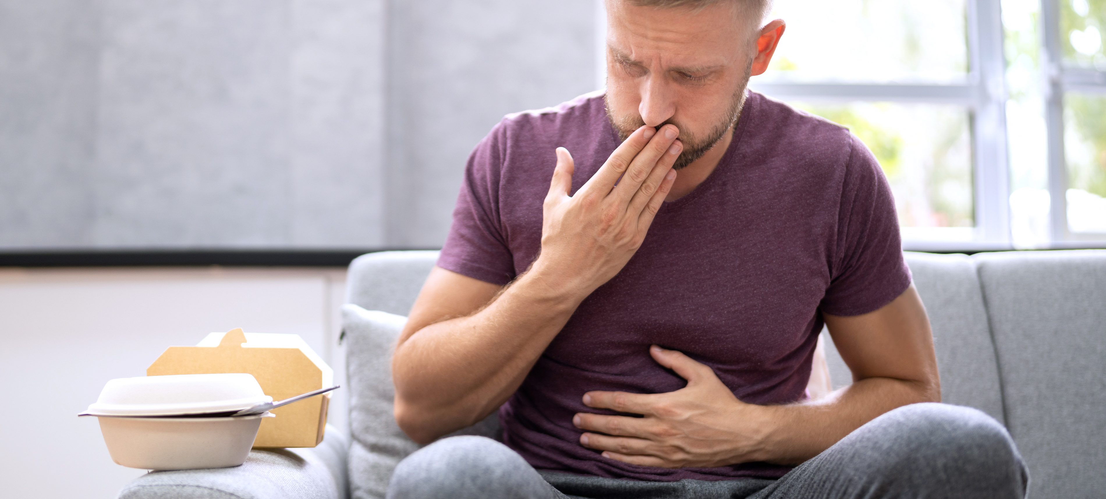 Ein junger Mann sitzt auf der Couch und hält sich eine Hand vor seinen Bauch und die andere Hand vor seinen Mund. Es sieht aus, als hätte er Sodbrennen und muss aufstoßen. | VIACTIV Krankenkasse