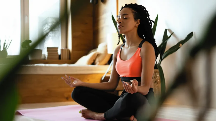 Eine junge Frau macht Yoga. Sie sitzt im Schneidersitz auf dem Fußboden und hat die Augen geschlossen. | VIACTIV Krankenkasse