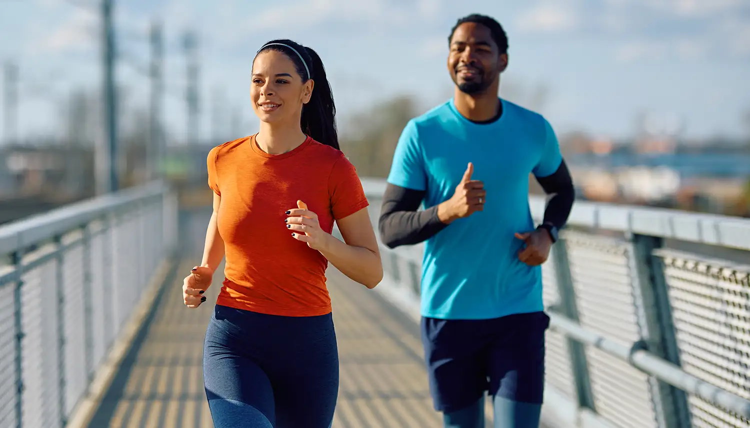 Zwei Personen joggen über eine Brücke. Die Sonne scheint und beide lächeln. | VIACTIV Krankenkasse