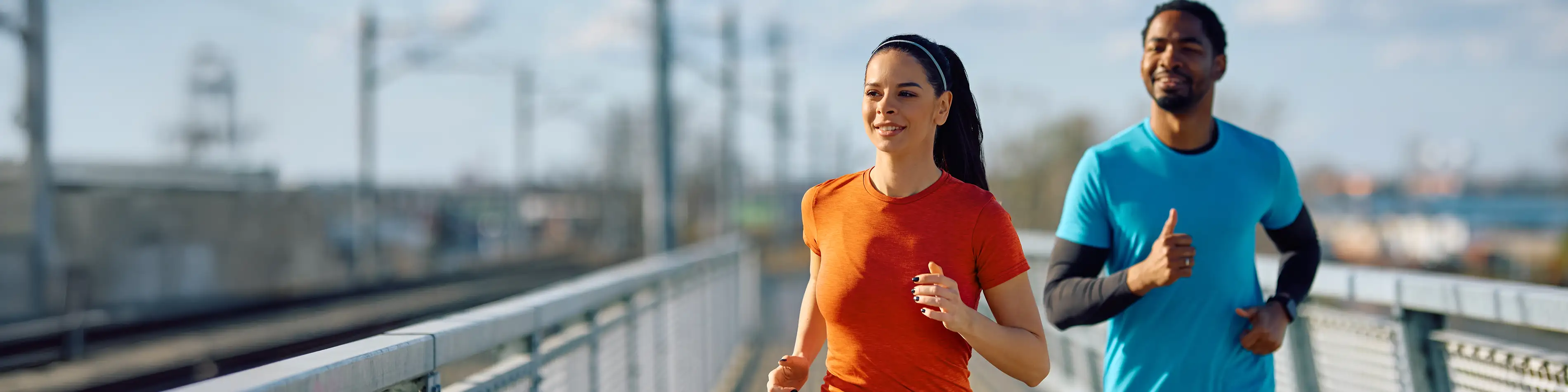 Zwei Personen joggen über eine Brücke. Die Sonne scheint und beide lächeln. | VIACTIV Krankenkasse