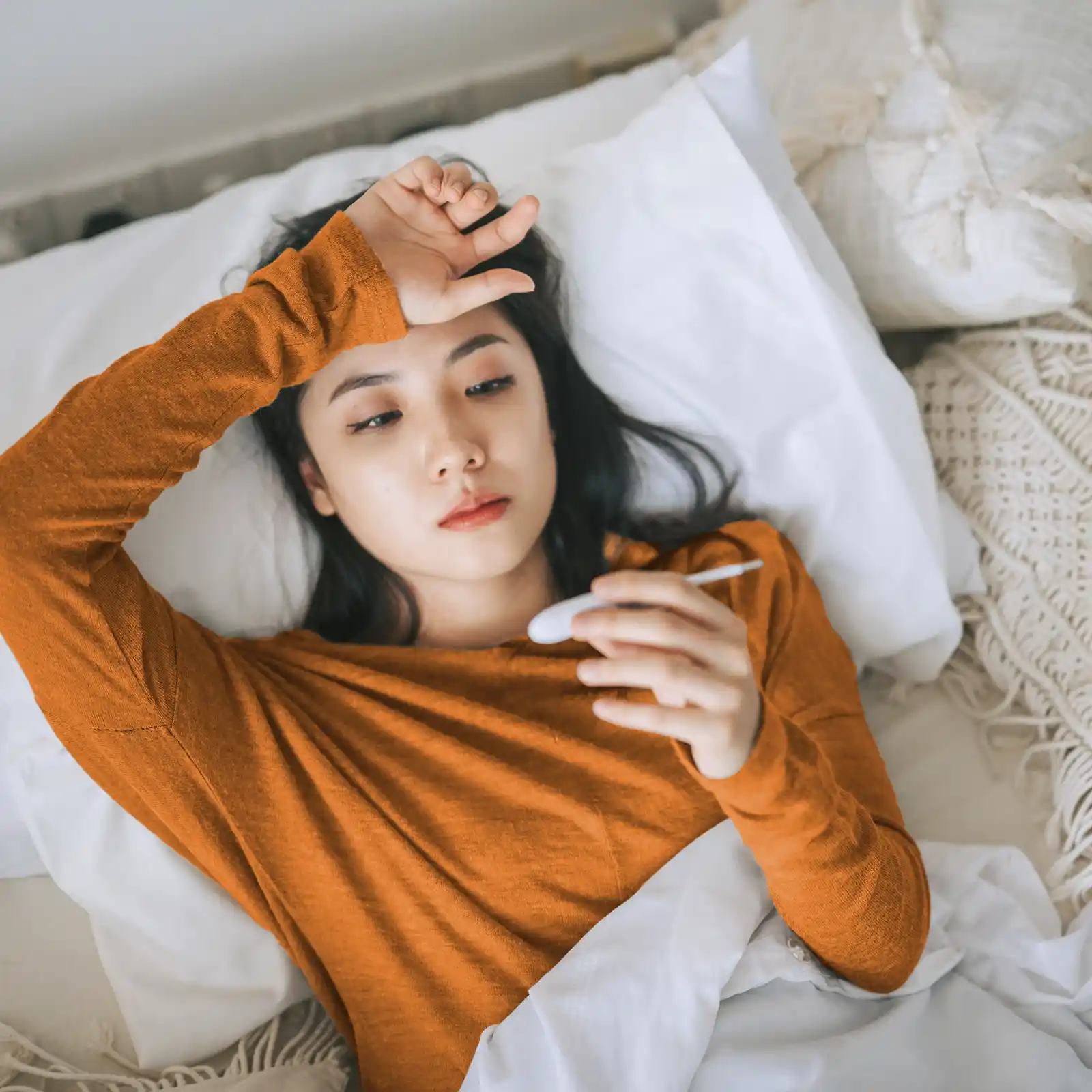 Eine junge Frau liegt im Bett und schaut auf ein Fieberthermometer. Ihr anderer Arm liegt auf ihrem Kopf. | VIACTIV Krankenkasse