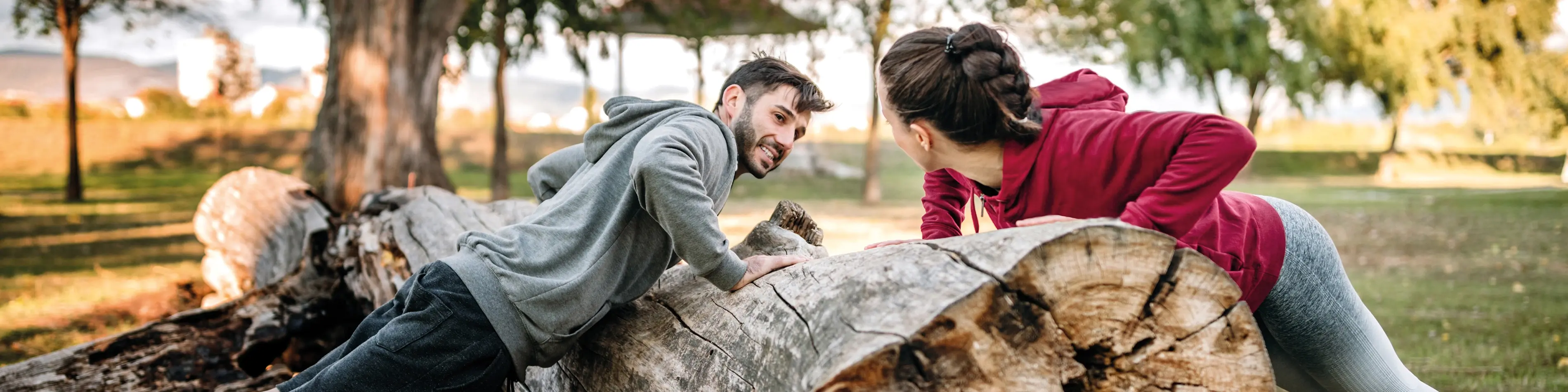 Junger Mann und junge Frau machen Outdoor Fitness an einem abgesägten Baum im Park bei Sonnenschein. | VIACTIV Krankenkasse