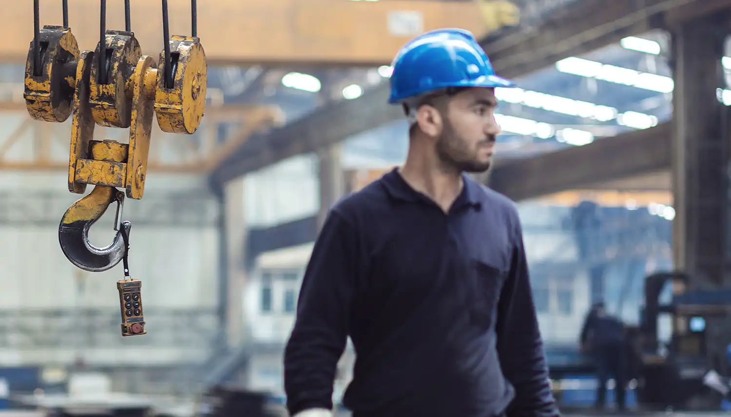 Ein männlicher Arbeiter mit Helm steht im Vordergrund und hinter ihm ist ein massiver Seilzug in einer Halle zu sehen. | VIACTIV Krankenkasse