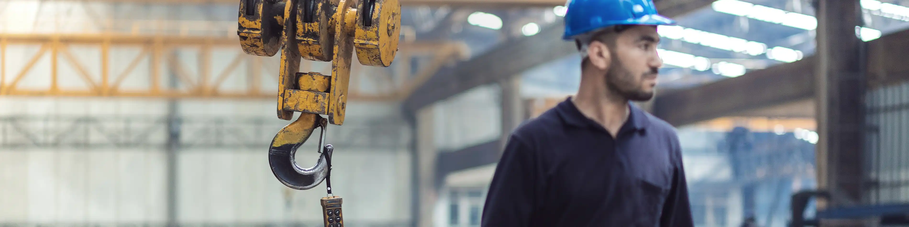 Ein männlicher Arbeiter mit Helm steht im Vordergrund und hinter ihm ist ein massiver Seilzug in einer Halle zu sehen. | VIACTIV Krankenkasse