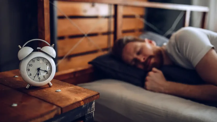 Ein Mann liegt schlafend im Bett. Im Vordergrund steht ein weißer großer Wecker mit den Zeigern auf 6:20 Uhr. | VIACTIV Krankenkasse