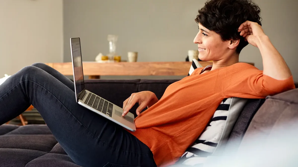 Eine Frau sitrzt gemütlich auf der Couch und bedient lächelnd einen Laptop auf ihrem Schoß. | VIACTIV Krankenkasse