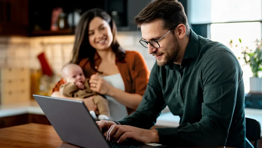 Ein Mann sitzt am Laptop und tippt etwas ein, während eine Frau im Hintergrund mit einem Baby auf dem Arm lächelt | VIACTIV Krankenkasse