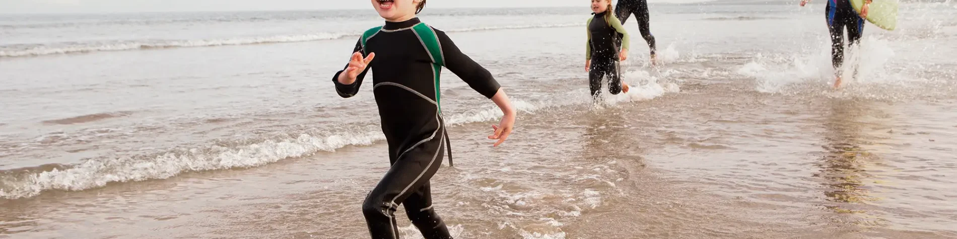 Eine vierköpfige Familie läuft in Neoprenanzügen am Strand entlang. | VIACTIV Krankenkasse
