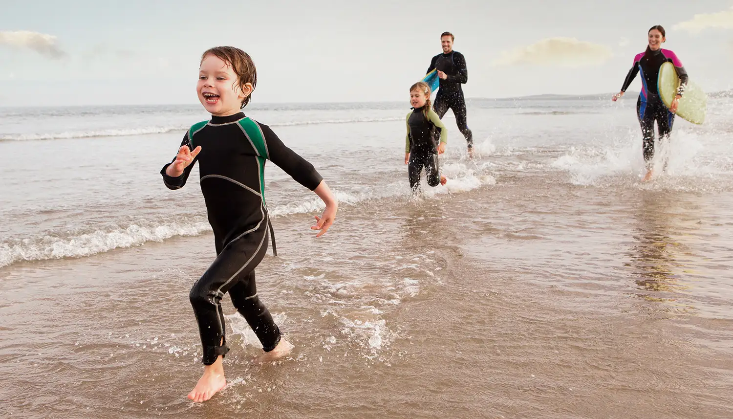 Eine vierköpfige Familie läuft in Neoprenanzügen am Strand entlang. | VIACTIV Krankenkasse