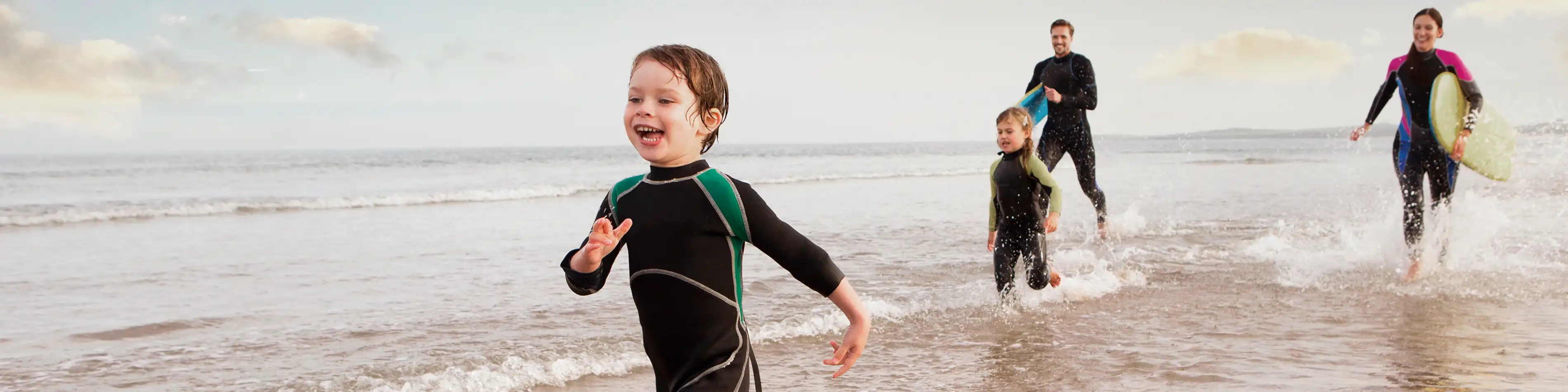 Eine vierköpfige Familie läuft in Neoprenanzügen am Strand entlang. | VIACTIV Krankenkasse