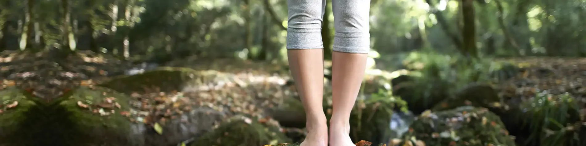 Eine Person steht mit nackten Füßen mitten im Wald bei Sonnenschein auf einem kleinen Hügel, der mit Moos bedeckt ist. | VIACTIV Krankenkasse