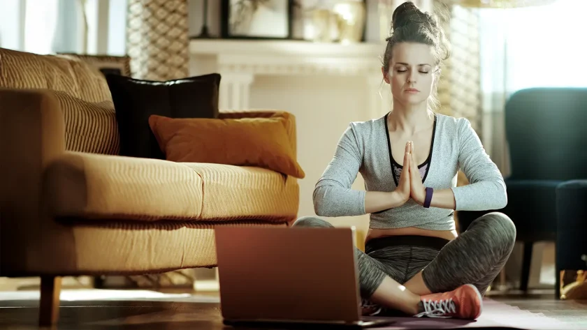 Eine junge Frau sitzt in einer Yogapose vor dem Laptop auf dem Boden in ihrem Wohnzimmer. Sie hat die Augen geschlossen. | VIACTIV Krankenkasse