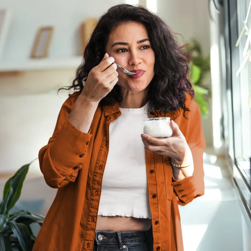 Eine junge Frau isst einen Joghurt. Dabei steht sie am Fenster und schaut raus. | VIACTIV Krankenkasse