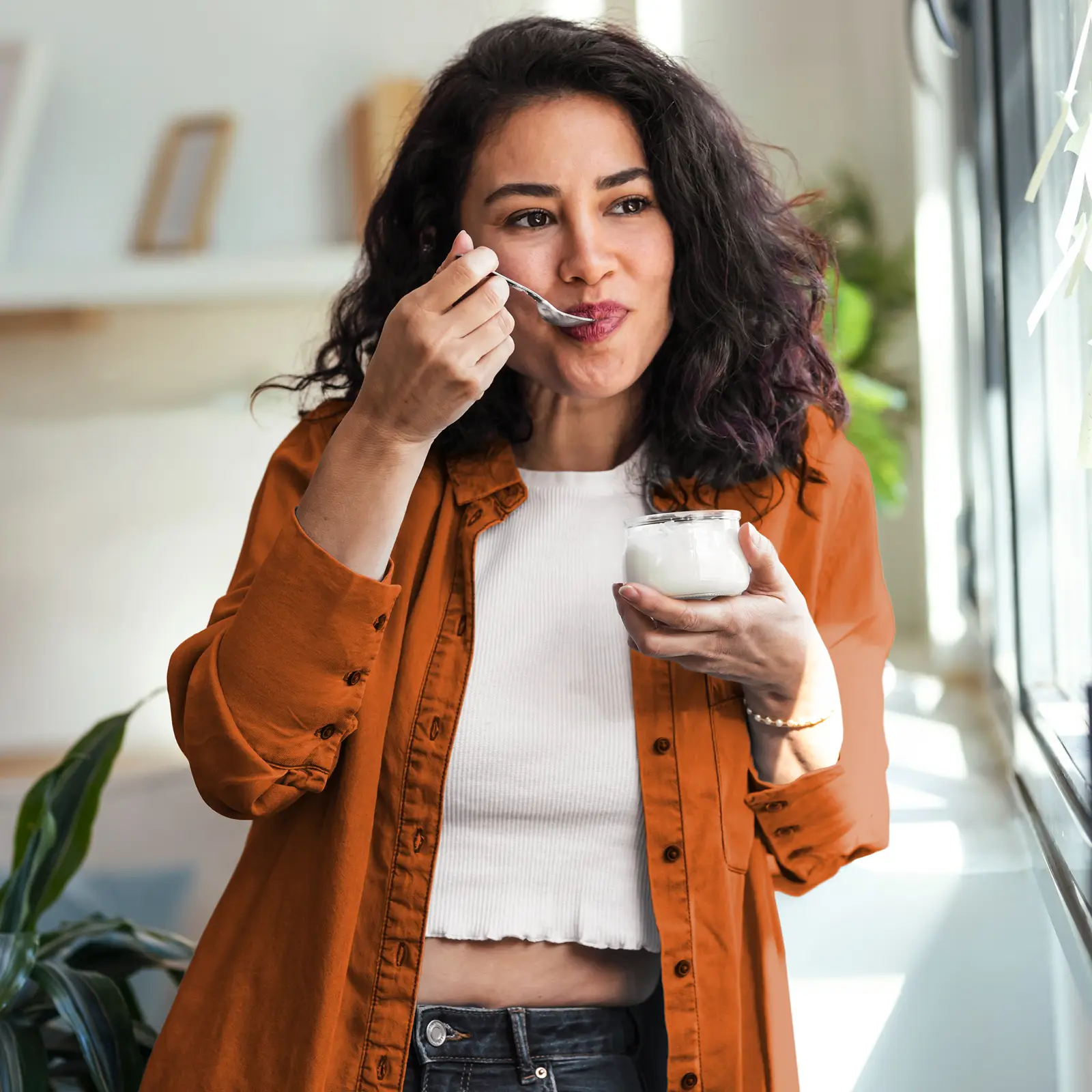 Eine junge Frau isst einen Joghurt. Dabei steht sie am Fenster und schaut raus. | VIACTIV Krankenkasse