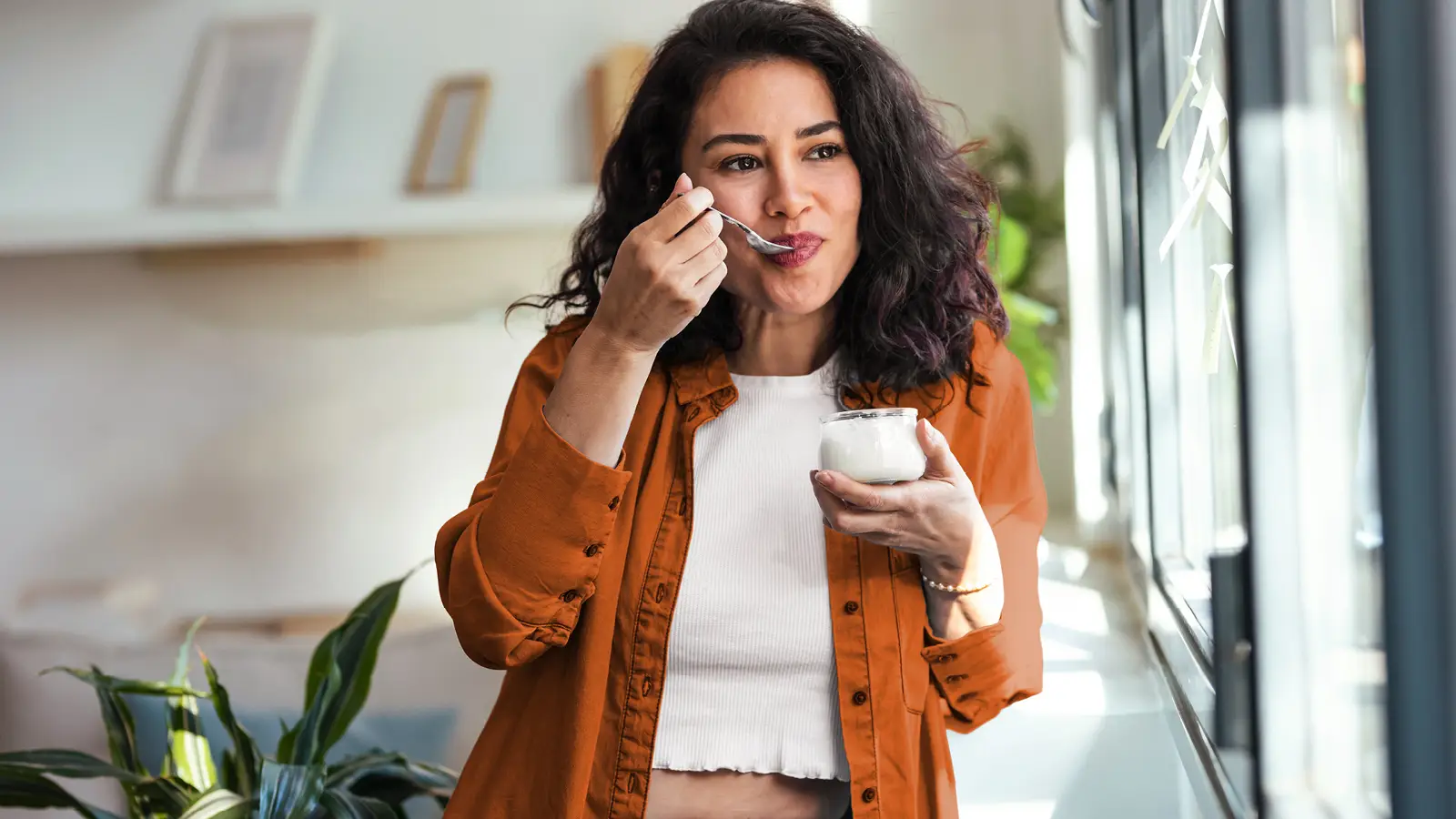 Eine junge Frau isst einen Joghurt. Dabei steht sie am Fenster und schaut raus. | VIACTIV Krankenkasse