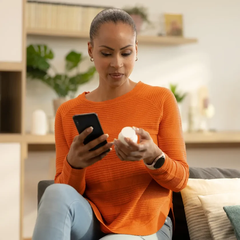 Eine junge Frau hält eine Medikamentenpackung in der Hand. In der anderen Hand hält sie ihr Smartphone. Sie liest den digitalen Beipackzettel. | VIACTIV Krankenkasse