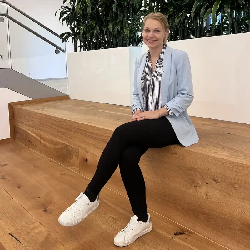 Auf dem Bild ist eine Mitarbeiterin der VIACTIV Krankenkasse zu sehen. Sie sitzt auf einer Treppe und lächelt. | VIACTIV Krankenkasse