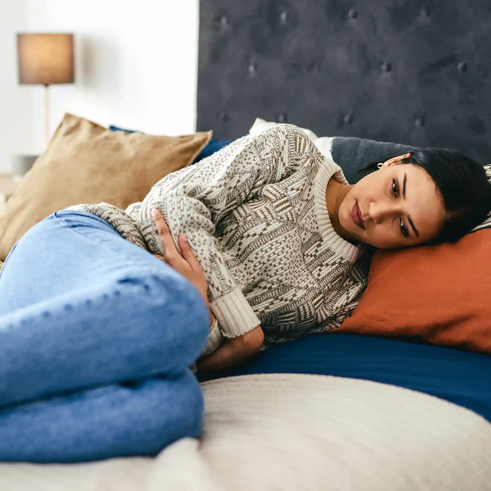 Eine junge Frau liegt auf dem Sofa und hält sich aufgrund von Unterleibschmerzen die Hände über den Bauch. | VIACTIV Krankenkasse