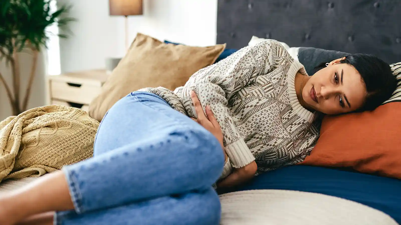 Eine junge Frau liegt auf dem Sofa und hält sich aufgrund von Unterleibschmerzen die Hände über den Bauch. | VIACTIV Krankenkasse