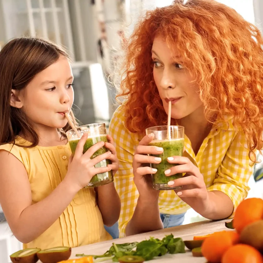 Eine junge Frau und ein kleines Mädchen trinken gemeinsam einen Smoothie. Vor ihnen auf dem Tisch liegt eine Portion Obst. | VIACTIV Krankenkasse