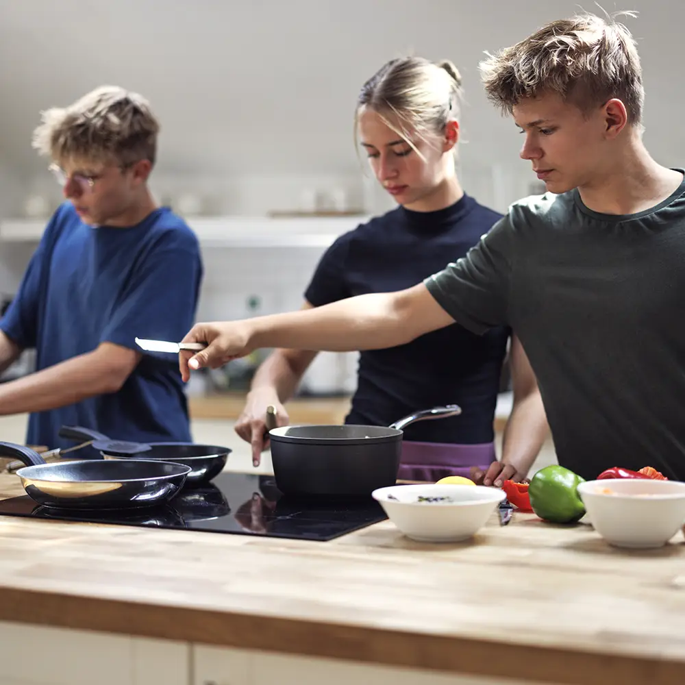 Auf dem Foto sind drei Jugendliche gemeinsam am kochen. Im Vordergrund ist ein Herd mit Töpfen und Pfannen zu sehen. | VIACTIV Krankenkasse