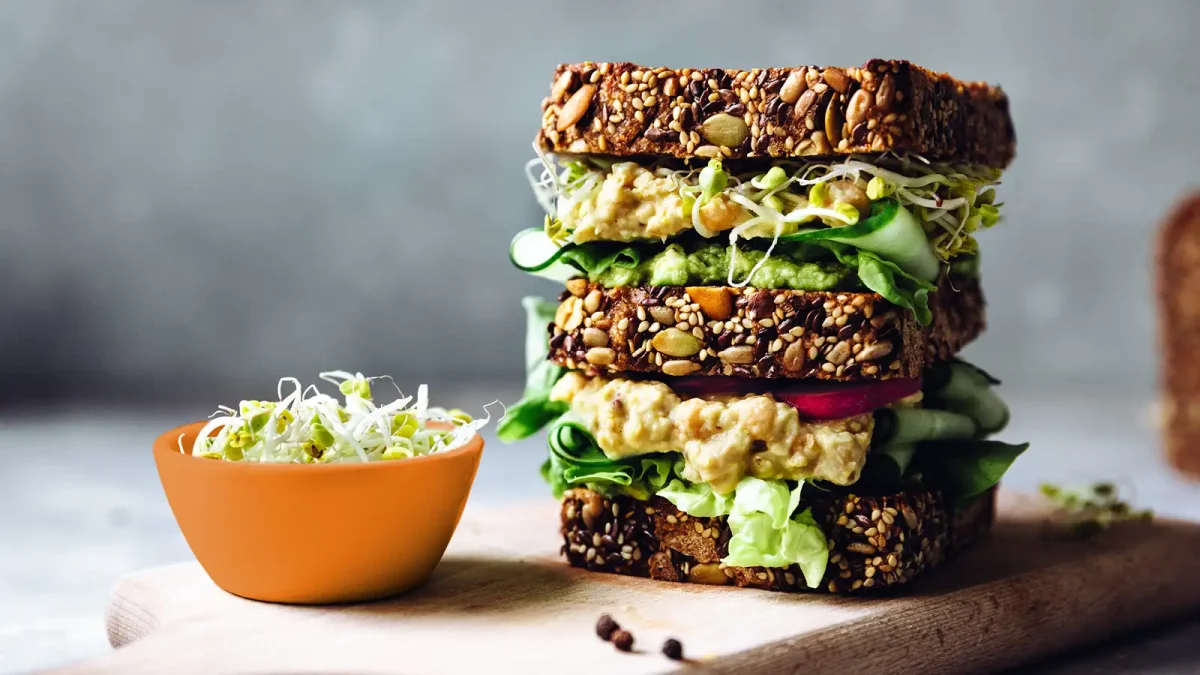 Ein mehrlagiges Sandwich aus Vollkornbrot und frischem Gemüse neben einer weißen Schale mit Salat. | VIACTIV Krankenkasse