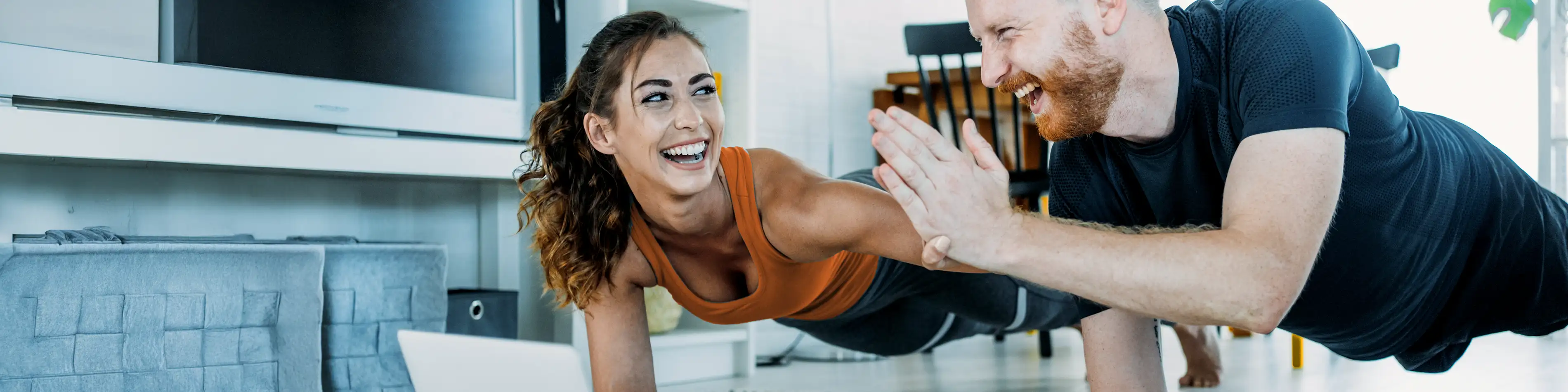 Zwei Personen machen nebeneinander die Fitnessübung Plank und klatschen sich dabei ab. Sie sind zu Hause in ihrem Wohnzimmer. | VIACTIV Krankenkasse