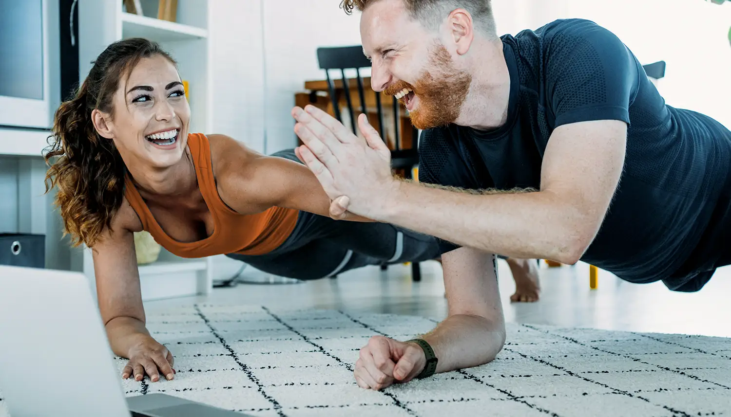 Zwei Personen machen nebeneinander die Fitnessübung Plank und klatschen sich dabei ab. Sie sind zu Hause in ihrem Wohnzimmer. | VIACTIV Krankenkasse