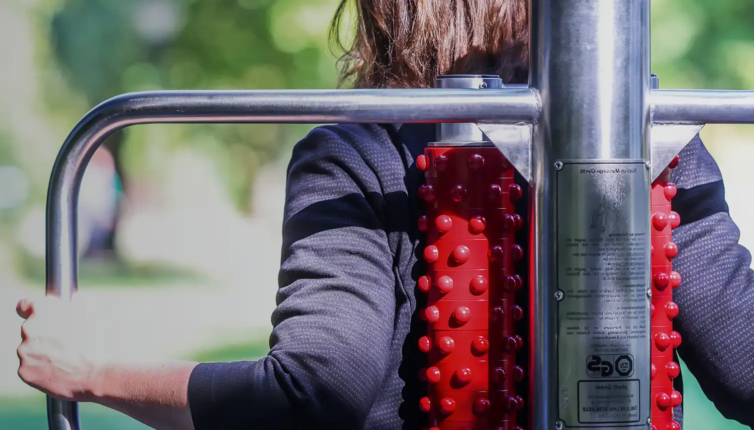 Eine Frau sitzt draußen bei Sonnenschein auf einem Parcour-Fitnessgerät mit dem Rücken zur Kamera gerichtet. | VIACTIV Krankenkasse