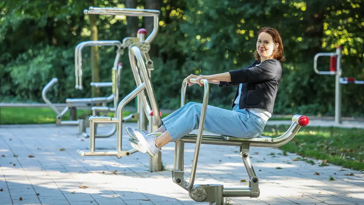 Eine Frau sitzt draußen bei Sonnenschein auf einem Parcour-Fitnessgerät mit Blick in die Kamera. | VIACTIV Krankenkasse