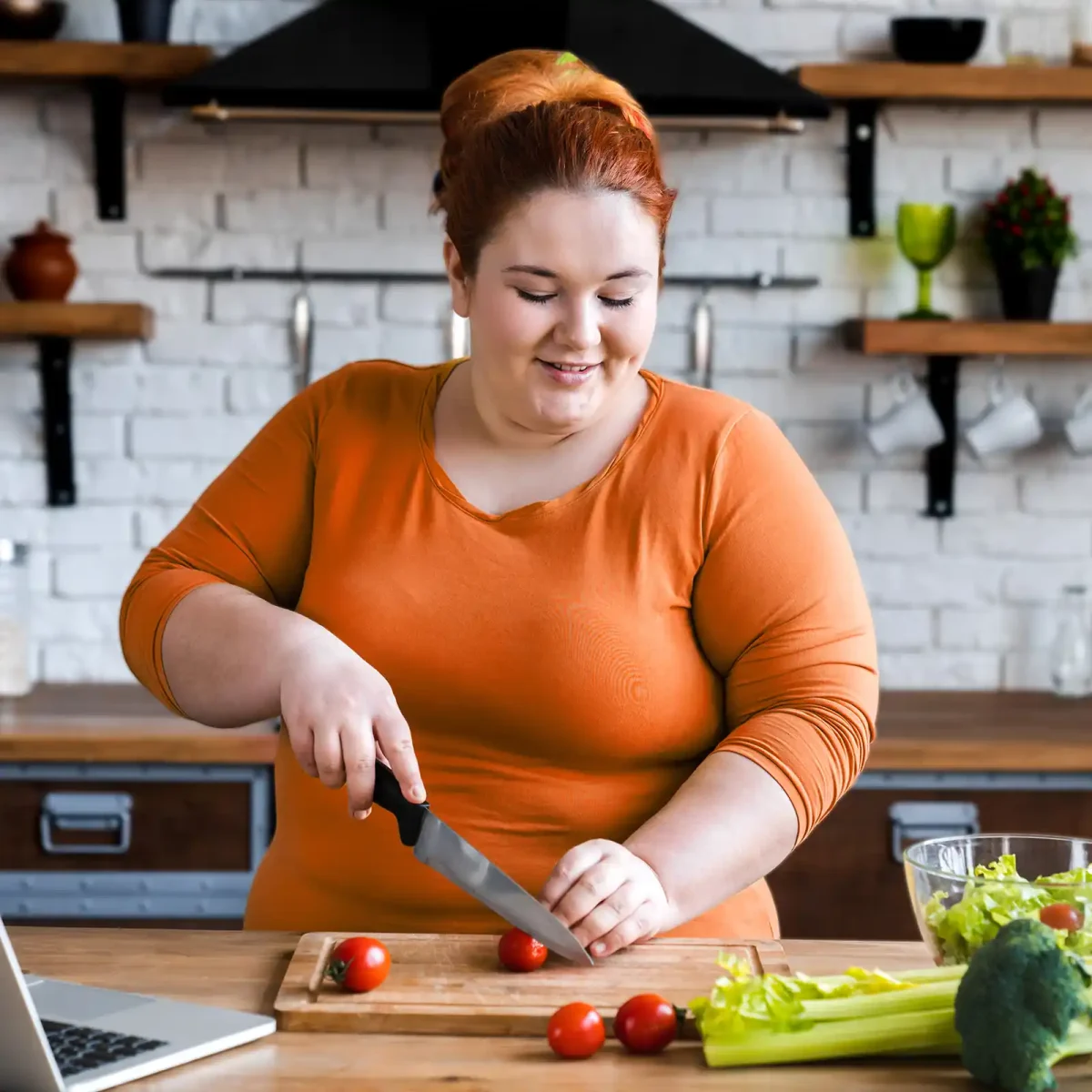 Eine junge Frau steht in der Küche, schneidet Gemüse und hat ihren Laptop aufgeklappt. | VIACTIV Krankenkasse