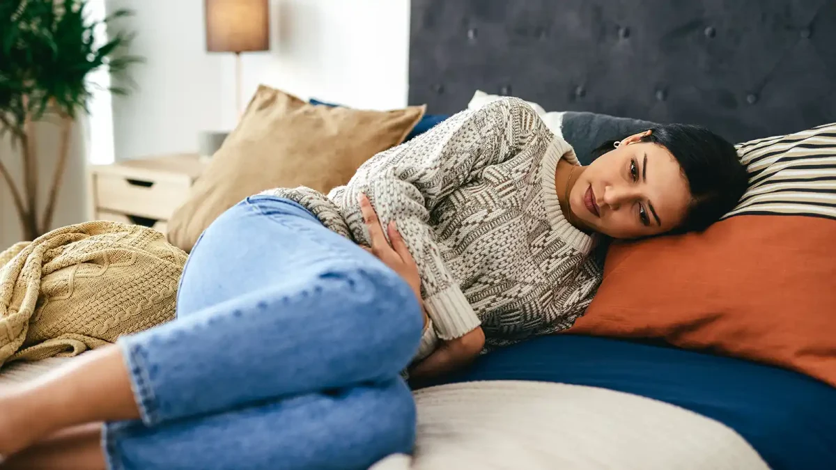 Eine junge Frau liegt auf dem Bett und hält ihre Arme um den Bauch. Sie hat Schmerzen. | VIACTIV Krankenkasse
