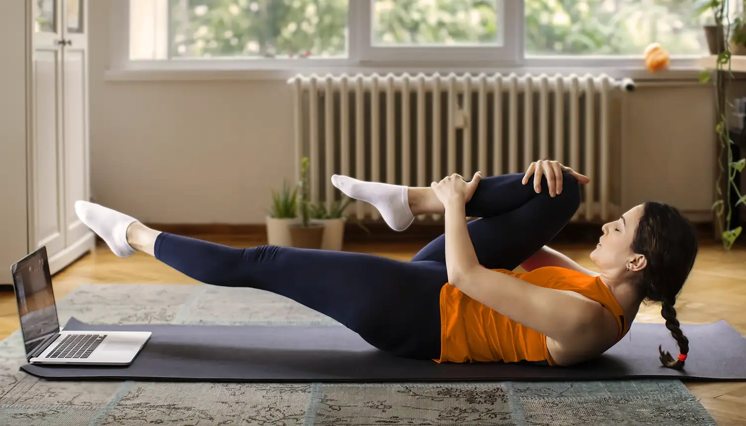 Eine Frau liegt in ihrem Wohnzimmer auf der Matte und macht eine Sportübung. Vor ihr steht ein Laptop, auf den sie blickt. | VIACTIV Krankenkasse