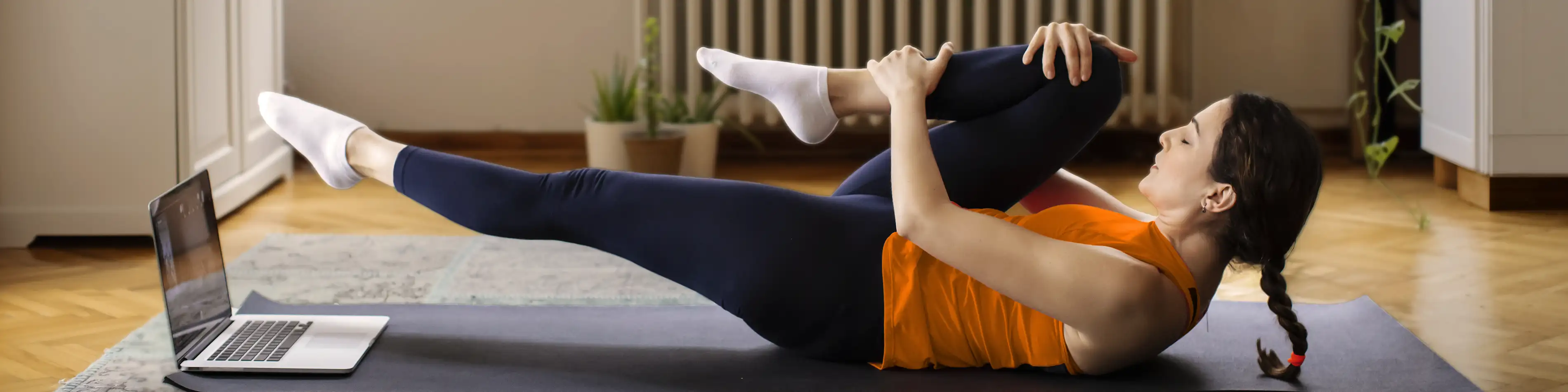 Eine Frau liegt in ihrem Wohnzimmer auf der Matte und macht eine Sportübung. Vor ihr steht ein Laptop, auf den sie blickt. | VIACTIV Krankenkasse