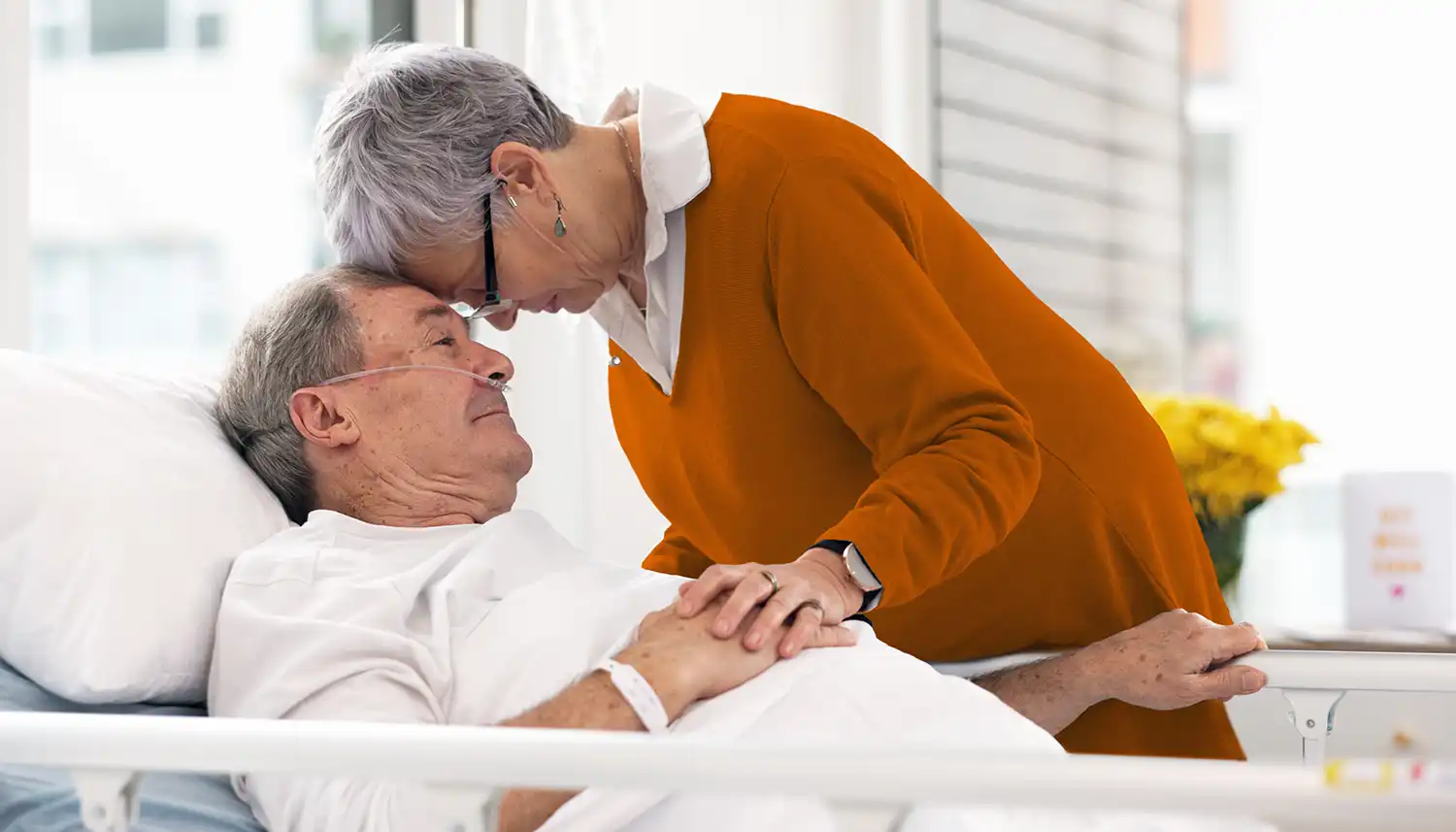 Ein älterer Mann mit Beatmungsgerät liegt im Krankenhaus. Eine Frau hält seine Hand und beugt sich zu ihm vor. | VIACTIV Krankenkasse
