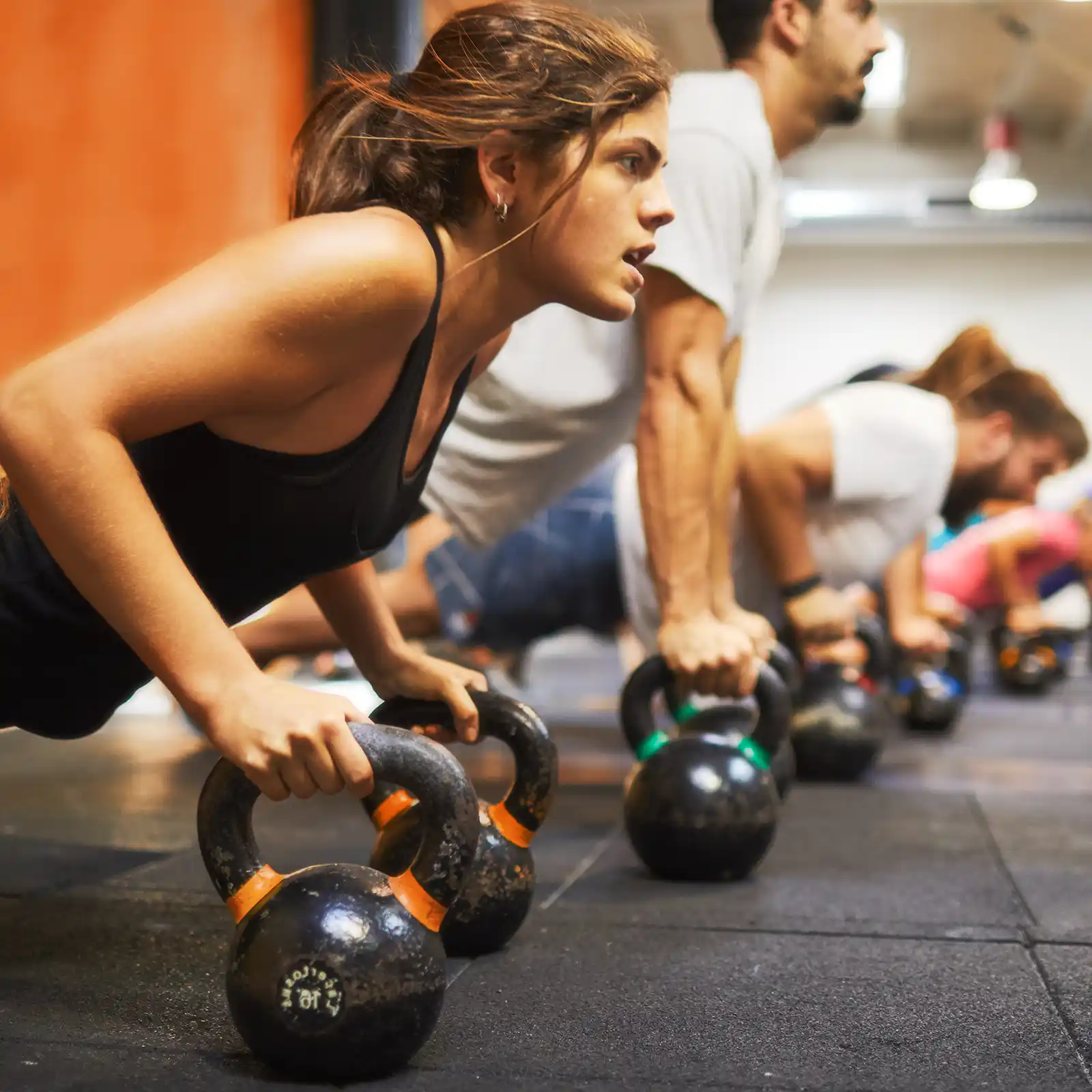 Eine Gruppe trainiert mit Kettlebells im Fitnessstudio. Die Frau im Vordergrund setz zum Liegestütz auf den Kettlebells an. | VIACTIV Krankenkasse