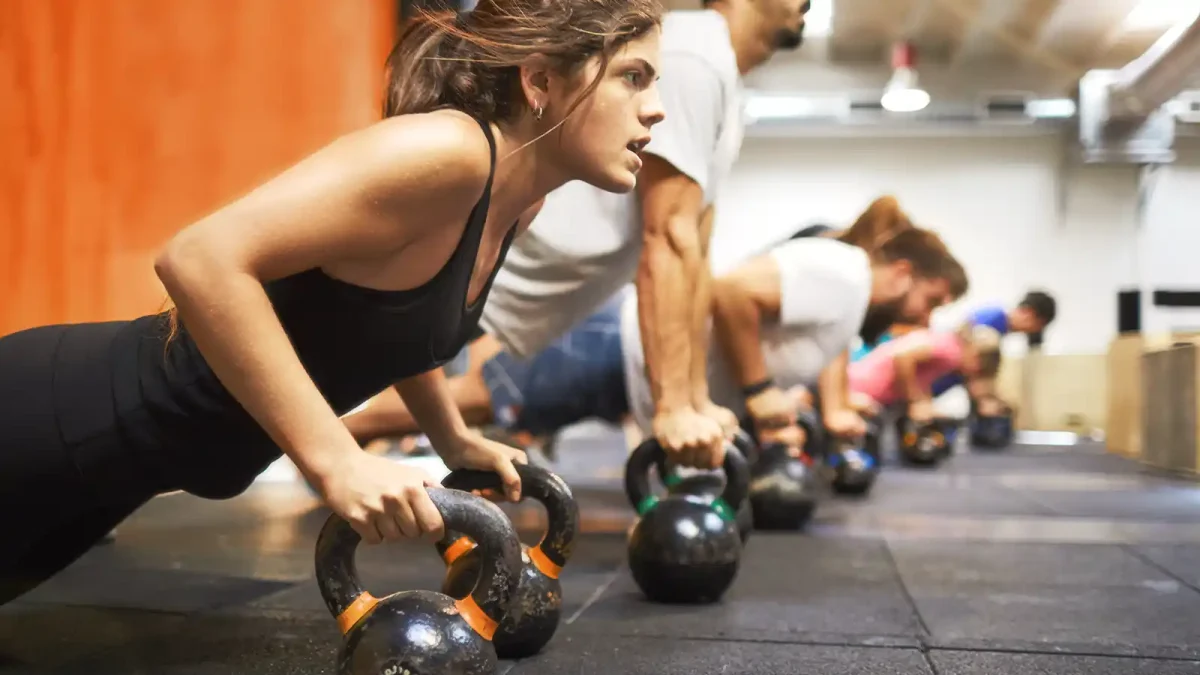 Eine Gruppe trainiert mit Kettlebells im Fitnessstudio. Die Frau im Vordergrund setzt zum Liegestütz auf den Kettlebells an. | VIACTIV Krankenkasse