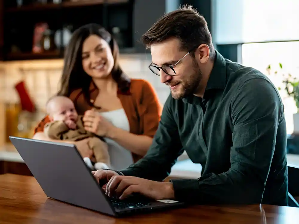 Im Hintergrund steht eine Frau hat einen Säugling auf dem Arm. Im Vordergrund steht ein Mann vor einem Laptop. | VIACTIV Krankenkasse