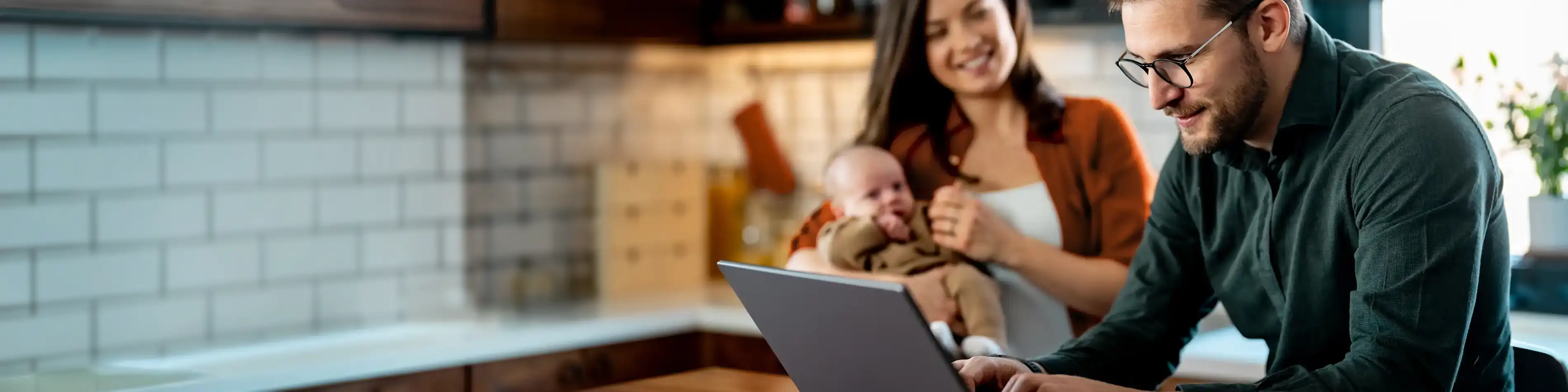 Im Hintergrund steht eine Frau hat einen Säugling auf dem Arm. Im Vordergrund steht ein Mann vor einem Laptop. | VIACTIV Krankenkasse
