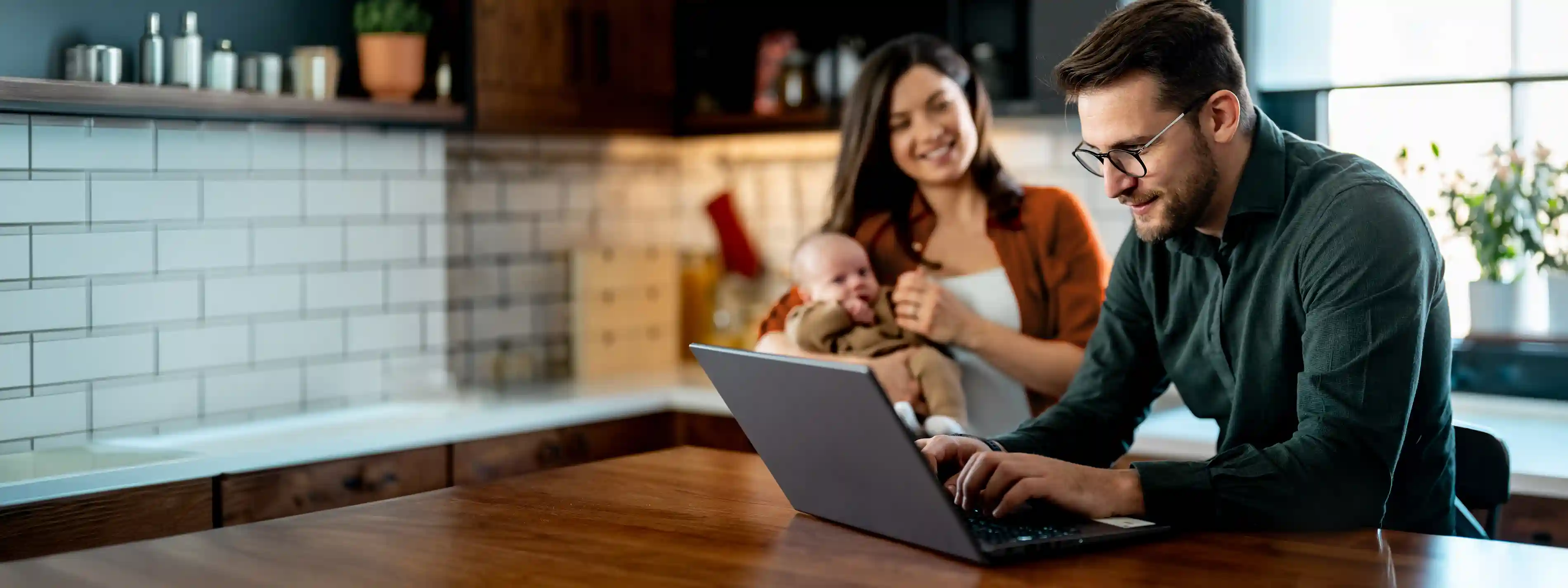 Im Hintergrund steht eine Frau hat einen Säugling auf dem Arm. Im Vordergrund steht ein Mann vor einem Laptop. | VIACTIV Krankenkasse