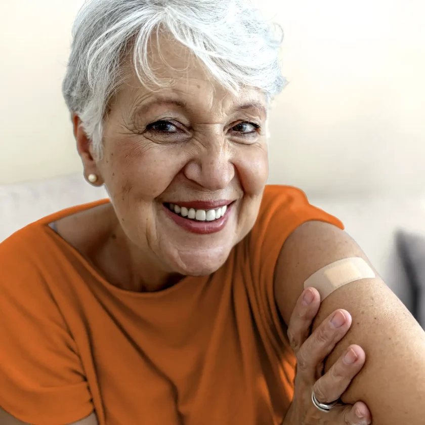 Eine Frau mit weißen kurzen Haaren lächelt. Man sieht ihre Zähne und sie trägt ein orangenes T-Shirt. Auf ihrem linken Oberarm ist ein Pflaster zu sehen. | VIACTIV Krankenkasse