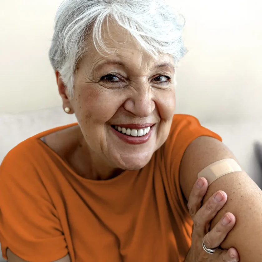 Eine Frau mit weißen kurzen Haaren lächelt. Man sieht ihre Zähne und sie trägt ein orangenes T-Shirt. Auf ihrem linken Oberarm ist ein Pflaster zu sehen. | VIACTIV Krankenkasse
