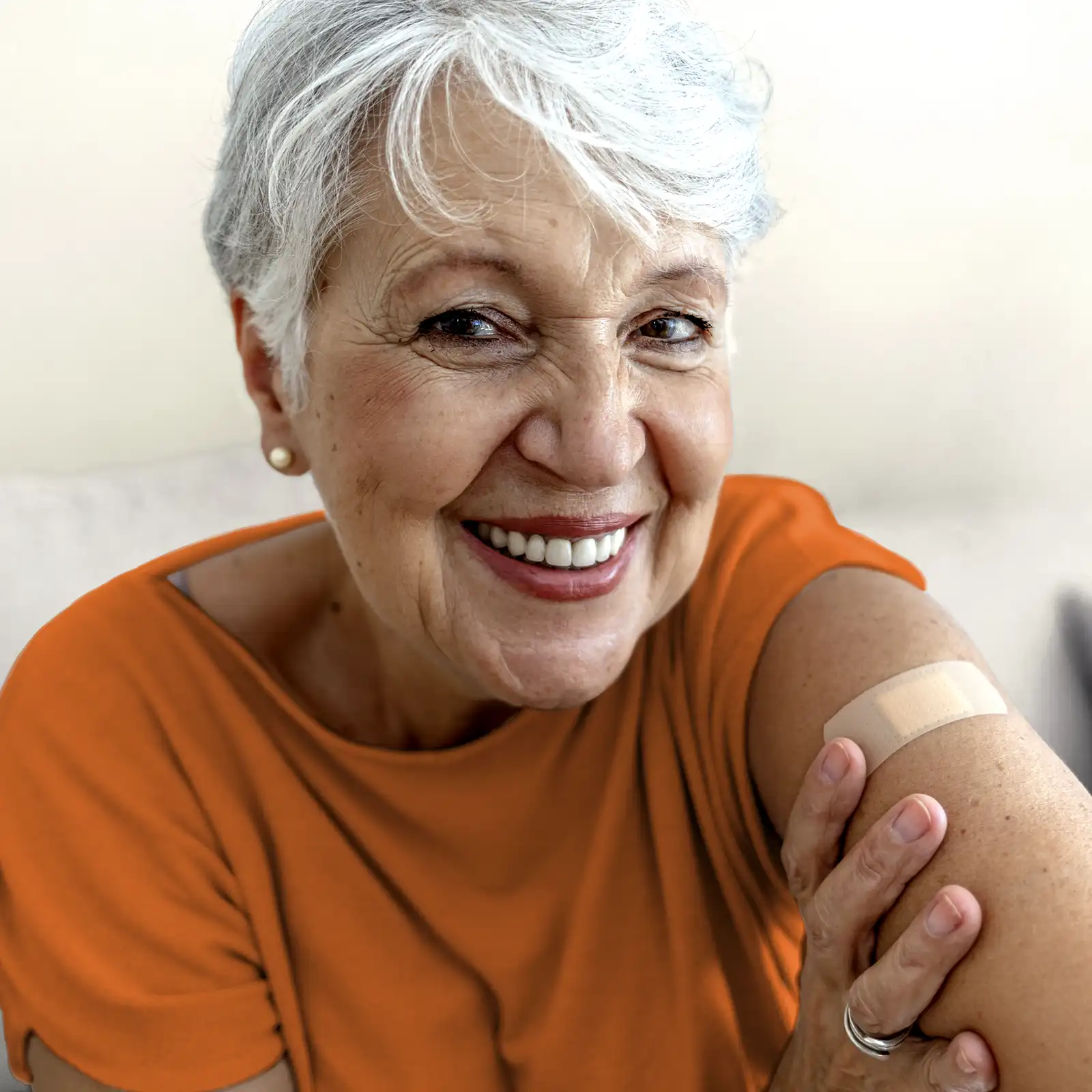 Eine Frau mit weißen kurzen Haaren lächelt. Man sieht ihre Zähne und sie trägt ein orangenes T-Shirt. Auf ihrem linken Oberarm ist ein Pflaster zu sehen. | VIACTIV Krankenkasse